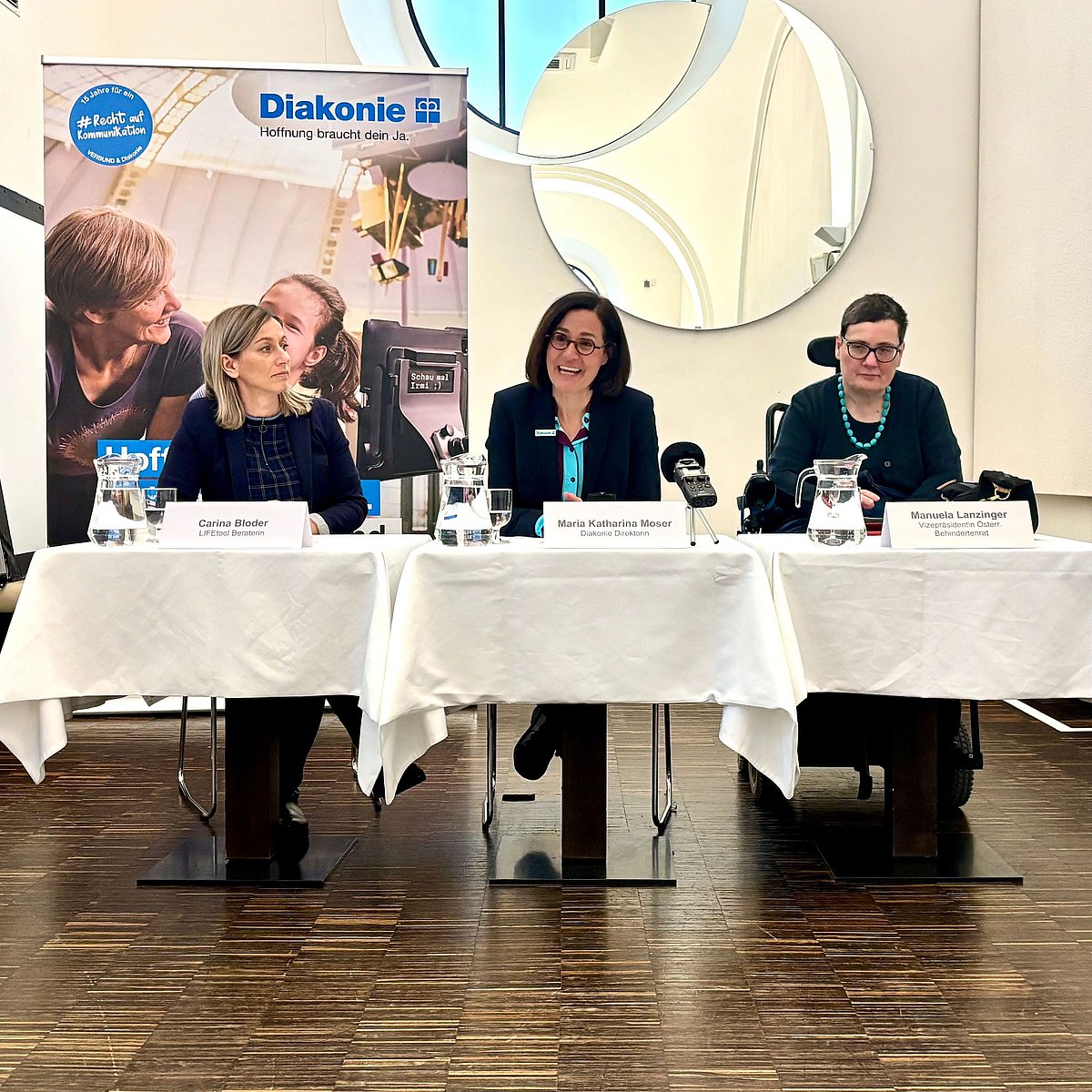 Drei Frauen sitzen an einem Tisch mit Mikrofonen bei einer Pressekonferenz. Dahinter ist ein Rollup mit den Logos von Diakonie und Verbund.